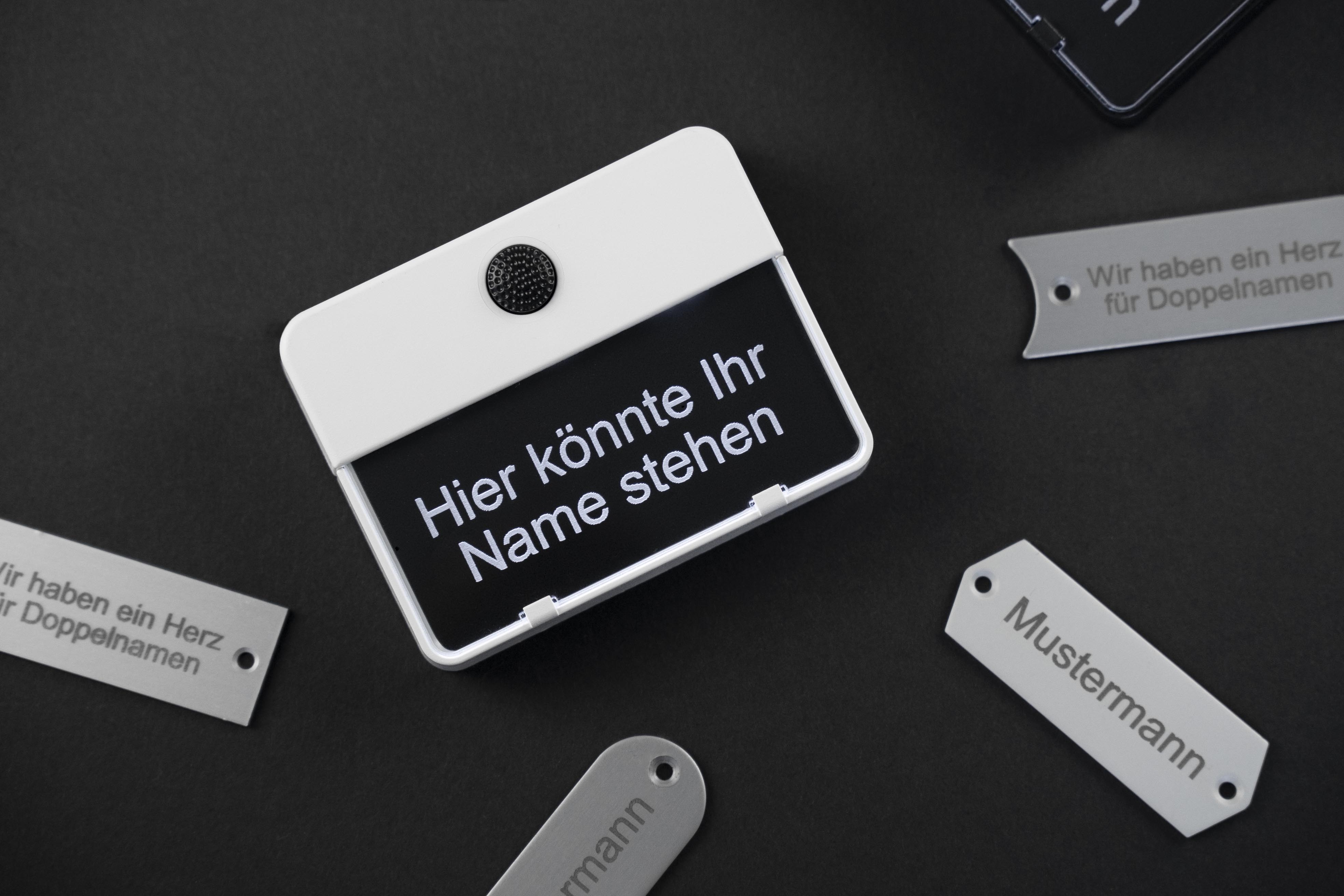 Ein Klingeltaster ETA 651 mit scharzem Gravierschild, auf dem "Hier könnte Ihr Name stehen" steht; darum liegen mehrere verschiedene Gravierschilder mit Schrift darauf; der Hintergrund ist dunkel