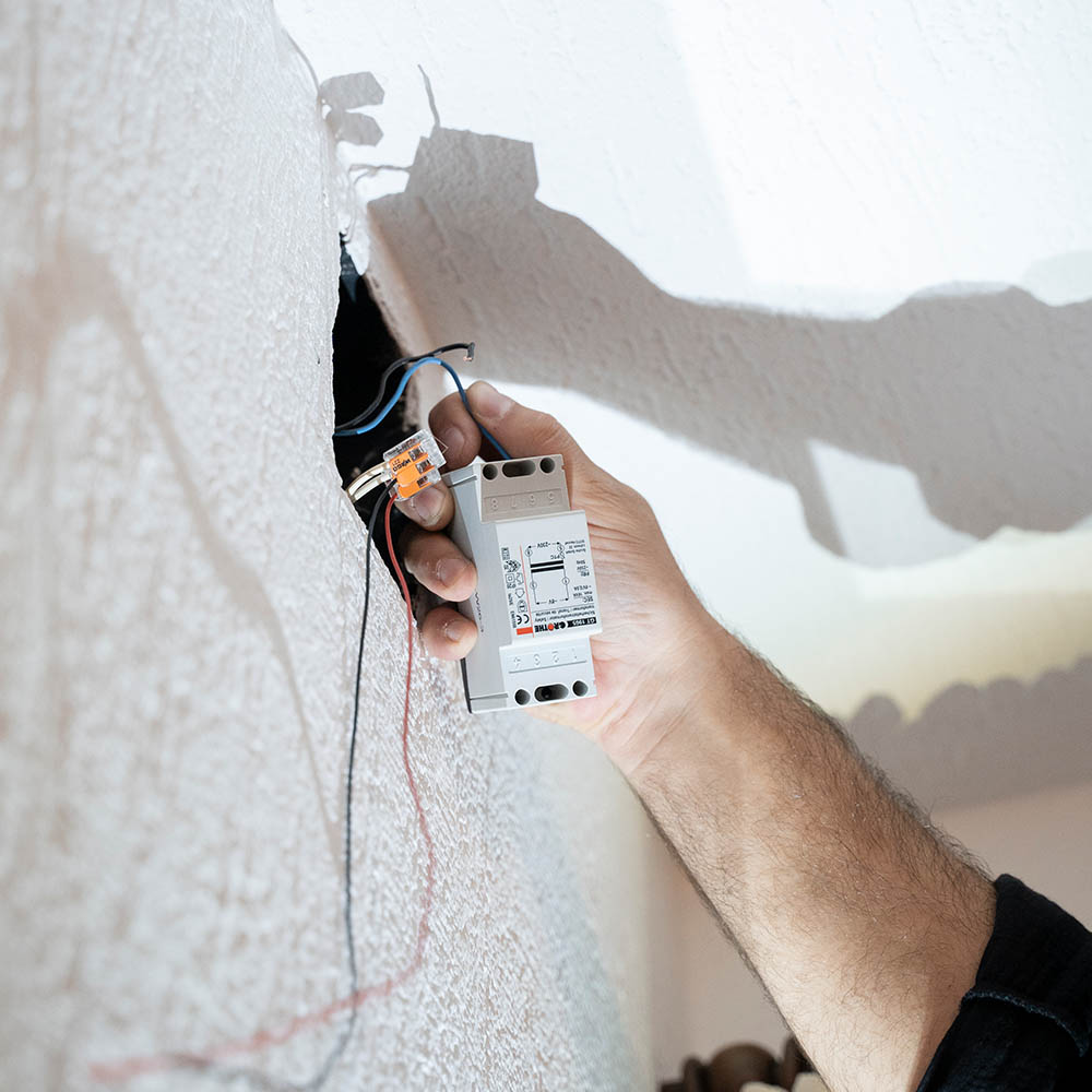 Jemand hält einen Trafo, der an Kabeln hängt, die aus einem Loch in der Wand ragen